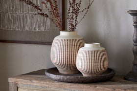 Two Perola Reactive Glaze Decorative Vases by nkuku—one tall, one short—sit on a round, dark wooden tray on a rustic table. Dried branches fill the taller vase; textured artwork hangs in the background.