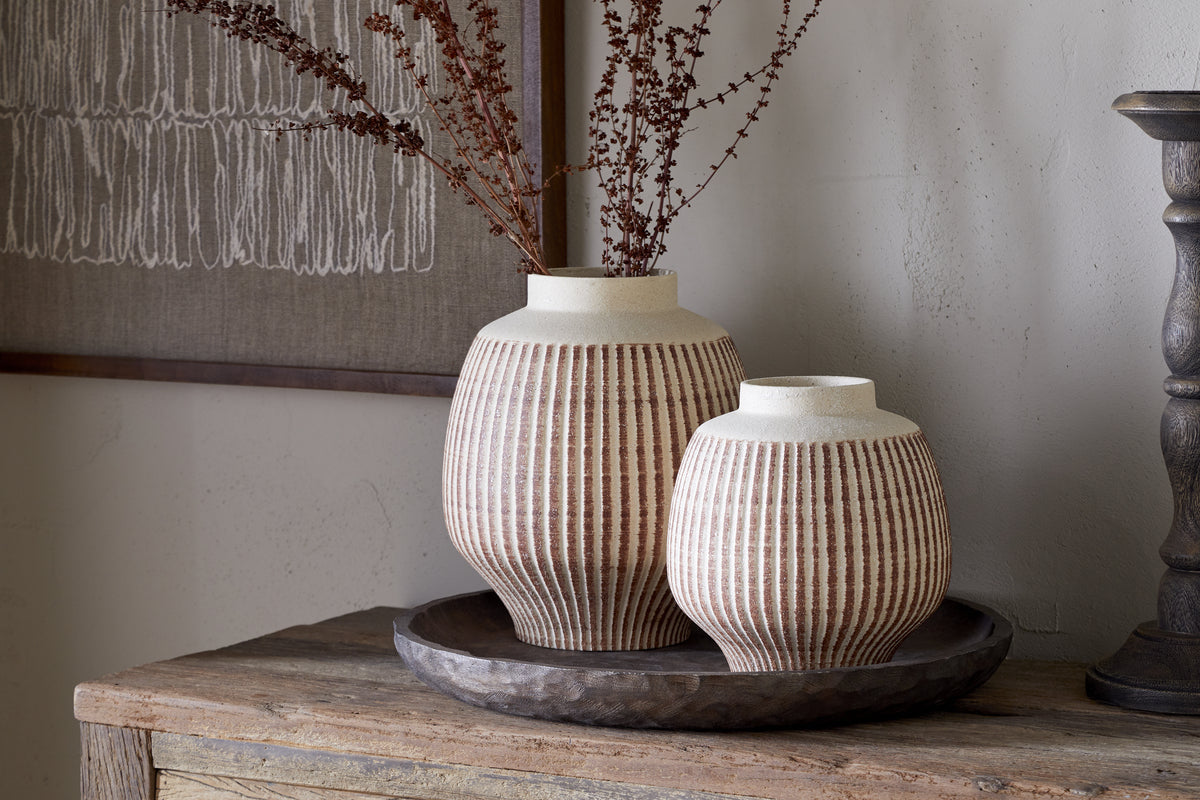 Two Perola Reactive Glaze Decorative Vases by nkuku—one tall, one short—sit on a round, dark wooden tray on a rustic table. Dried branches fill the taller vase; textured artwork hangs in the background.