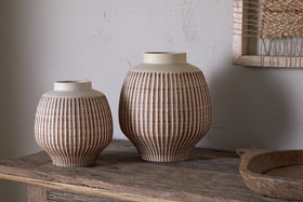 The nkuku Perola Reactive Glaze Decorative Vases in natural & rust, one small and one large with vertical ribbing, sit on a rustic table near a wooden tray and woven wall art against a light textured wall.