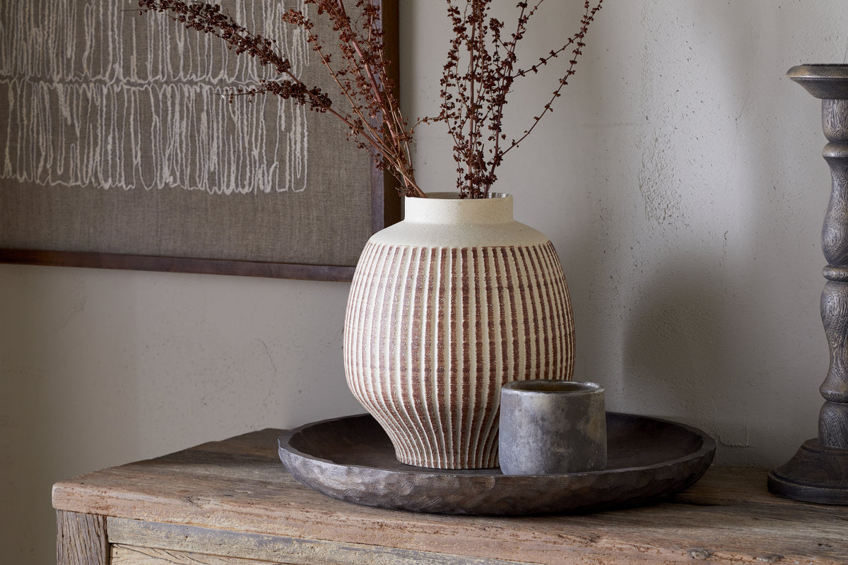 The nkuku Perola Reactive Glaze Decorative Vase in Natural & Rust with dried brown branches sits on a round wooden tray beside a gray candle atop a rustic wooden surface, backed by a textured wall and framed abstract art.