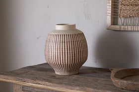 The nkuku Perola Reactive Glaze Decorative Vase in Natural & Rust sits on a rustic wooden table. Its round body tapers to a wide opening, set against a textured light gray wall with part of a framed woven artwork visible behind.