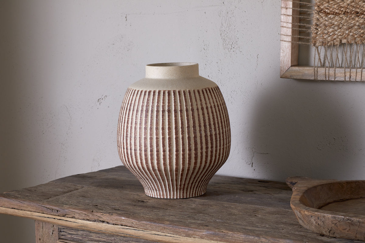 The nkuku Perola Reactive Glaze Decorative Vase in Natural & Rust sits on a rustic wooden table. Its round body tapers to a wide opening, set against a textured light gray wall with part of a framed woven artwork visible behind.