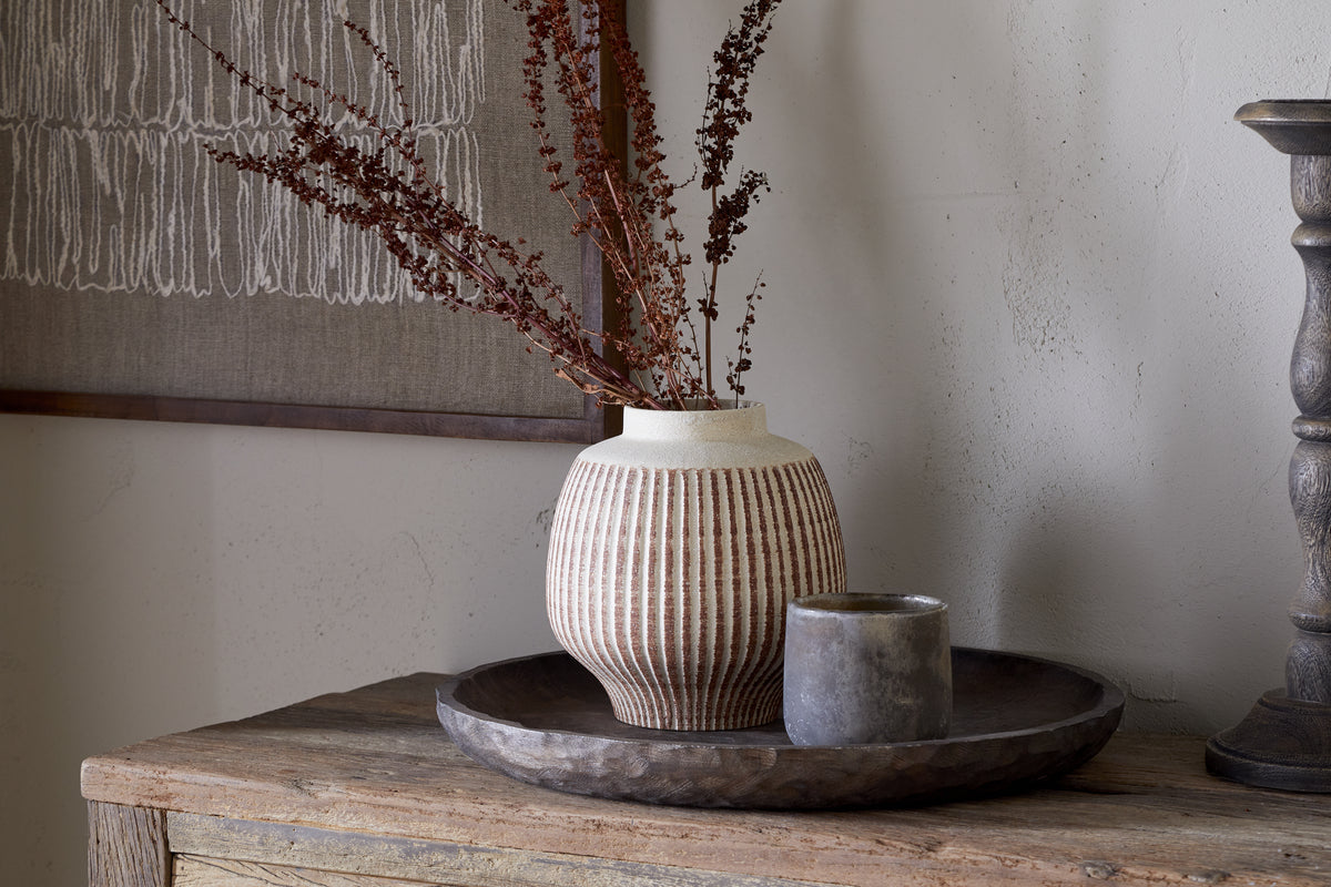 The nkuku Perola Reactive Glaze Decorative Vase in Natural & Rust holds dried brown branches on a round rustic wooden tray beside a small gray cup, with textile art hanging on a light wall in the background.