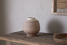 The nkuku Perola Reactive Glaze Decorative Vase in Natural & Rust, with parallel vertical brown and beige stripes, sits on a rustic wood table against a textured light wall and the edge of a framed woven artwork.