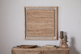The nkuku Pemali Woven Jute Wall Art - Natural is displayed above a rustic wooden sideboard with wooden candlesticks holding gray candles, a shallow tray, and a book for cozy decor against a white wall.