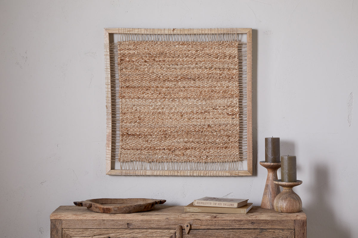 The nkuku Pemali Woven Jute Wall Art - Natural is displayed above a rustic wooden sideboard with wooden candlesticks holding gray candles, a shallow tray, and a book for cozy decor against a white wall.