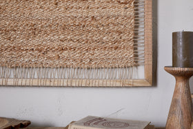 A close-up of the nkuku Pemali Woven Jute Wall Art - Natural, crafted with thick jute fibers and framed in mango wood, displayed beside a dark pillar candle on a wooden stand and a closed book on a light surface.
