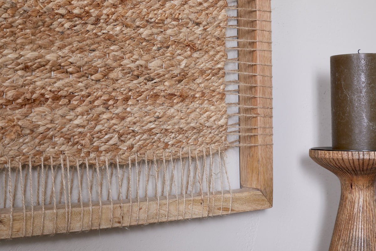 Close-up of the Pemali Woven Jute Wall Art - Natural by nkuku, featuring textured jute fibers in a mango wood frame beside a large brown candle on a wooden stand—ideal for adding rustic charm to any light-toned space.