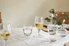 A simple table setting features nkuku’s Pani Wine Glass - Clear (Set of 4) filled with white wine and water on a light tablecloth. A eucalyptus-filled vase adds freshness, and woven chairs in the background create an inviting, everyday dining vibe.