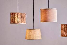 Four pendant lights with nkuku's Dia Jute Lampshade in natural hang from black cords against a light grey wall. Each shade features woven jute and wooden beads, adding textured, cozy ambient lighting.