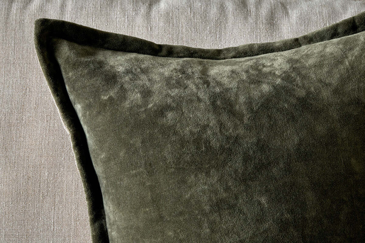 A close-up of the nkuku Pilani Velvet & Linen Cushion Cover in Moss, showcasing its soft, plush velvet texture and piped edges as it rests on a light gray fabric surface with lighting highlighting its subtle sheen.