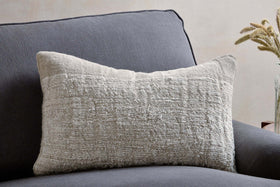 The nkuku Puli Recycled Linen Cushion Cover in Natural, with its soft textured beige linen, rests on a dark gray armchair. Light walls and a glass vase with dried grasses are partly visible in the background.