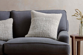 A dark gray upholstered sofa features two nkuku Puli Recycled Linen Cushion Covers in Natural. Next to it stands a wooden side table with a glass vase of dried grasses, set against a subtly textured, light-colored wall.