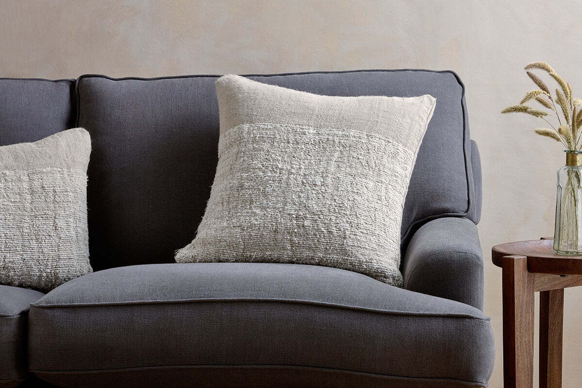 A dark gray upholstered sofa features two nkuku Puli Recycled Linen Cushion Covers in Natural. Next to it stands a wooden side table with a glass vase of dried grasses, set against a subtly textured, light-colored wall.