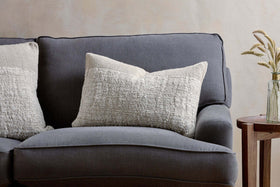 A dark gray fabric sofa with two large nkuku Puli Recycled Linen Cushion Covers in natural sits beside a small wooden side table topped with a glass vase of dried grasses, set against a light beige wall.