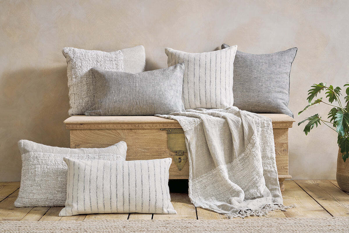 A wooden chest is topped with five nkuku Puli Recycled Linen Cushion Covers in natural, a light woven throw blanket, and two pillows on the floor. A leafy plant sits to the right against a beige wall and wooden floor for a cozy, minimalist vibe.