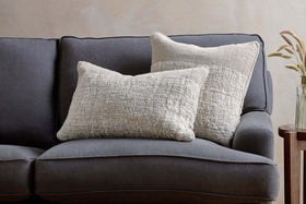 A dark gray upholstered couch features two nkuku Puli Recycled Linen Cushion Covers in natural, arranged against the backrest. On the right, a small wooden side table holds a glass vase with dried grasses. The wall behind is light and neutral.