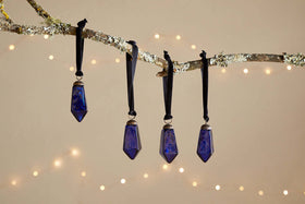 Four small, faceted indigo Ratnam Baubles by nkuku hang from black ribbons on a lichen-covered branch. Warm white fairy lights glow against a soft beige background, creating a festive, cozy scene. Set of 4 included.