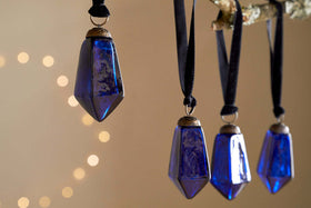 Four Ratnam Baubles - Indigo - Small by nkuku, with faceted dark blue glass and antique finish, hang from black cords on a branch. Soft yellow string lights create a cozy, festive atmosphere that highlights the ornaments.