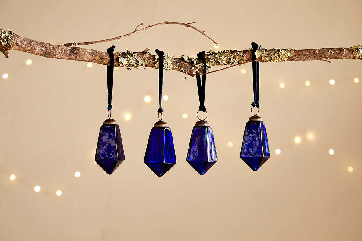 Four Ratnam Baubles - Indigo - Large (Set of 4) by nkuku, featuring geometric shapes and an antique glass finish, hang from black ribbons on a lichen branch. Soft fairy lights glow behind, evoking classic Christmas charm.