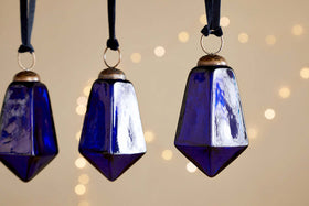 Four large, faceted indigo Ratnam Baubles by nkuku hang from black ribbons against a beige background with warm white lights, bringing an elegant, festive accent to classic Christmas decor.