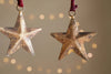 The Roshani Star Baubles - Antique Brass (Set of 4) by nkuku feature rustic, gold-toned stars with a textured, weathered finish. Hanging from red strings, they create a festive holiday feel against soft beige backgrounds.