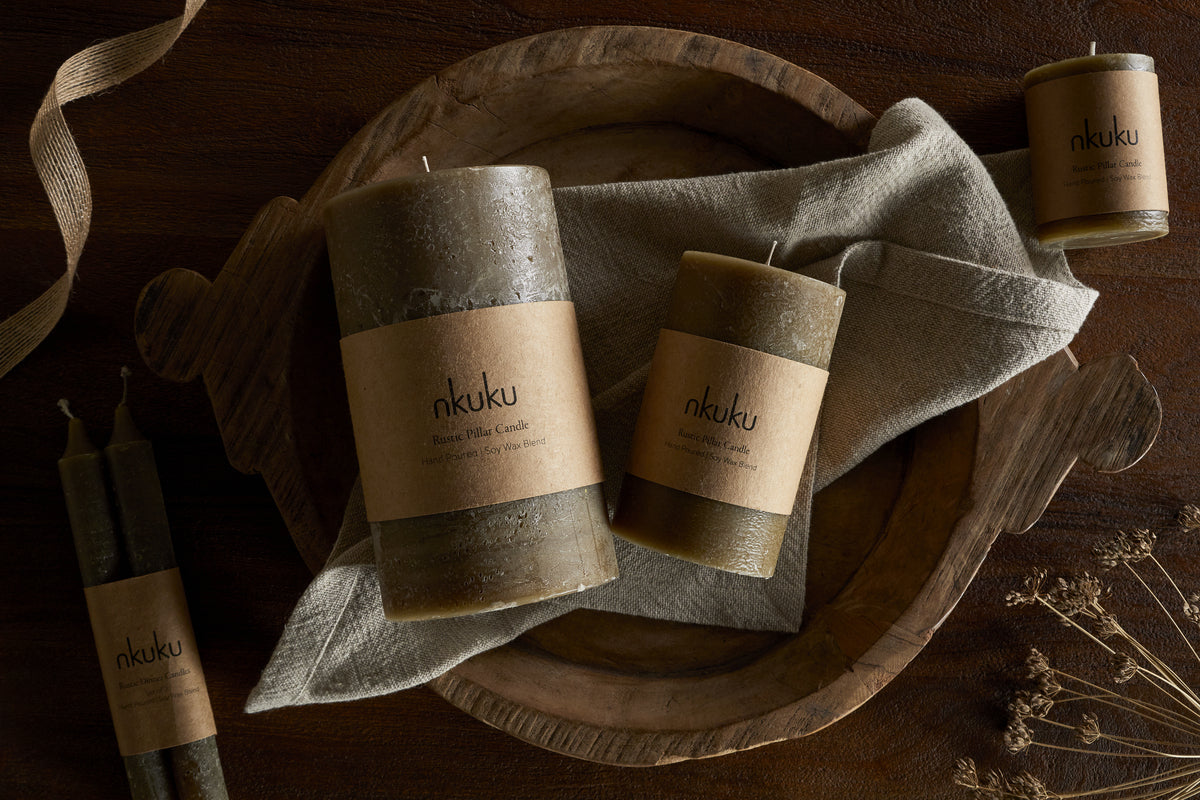 A rustic wooden tray holds three nkuku Rustic Soy Blend Mini Pillar Candles in Olive on a folded linen cloth, accented by two tapered candles and dried flowers atop a dark wooden surface.