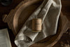 A Rustic Soy Blend Mini Pillar Candle - Olive by nkuku sits on a light beige cloth in a rustic wooden bowl, with part of an open book in the bottom left corner atop a dark wood surface.