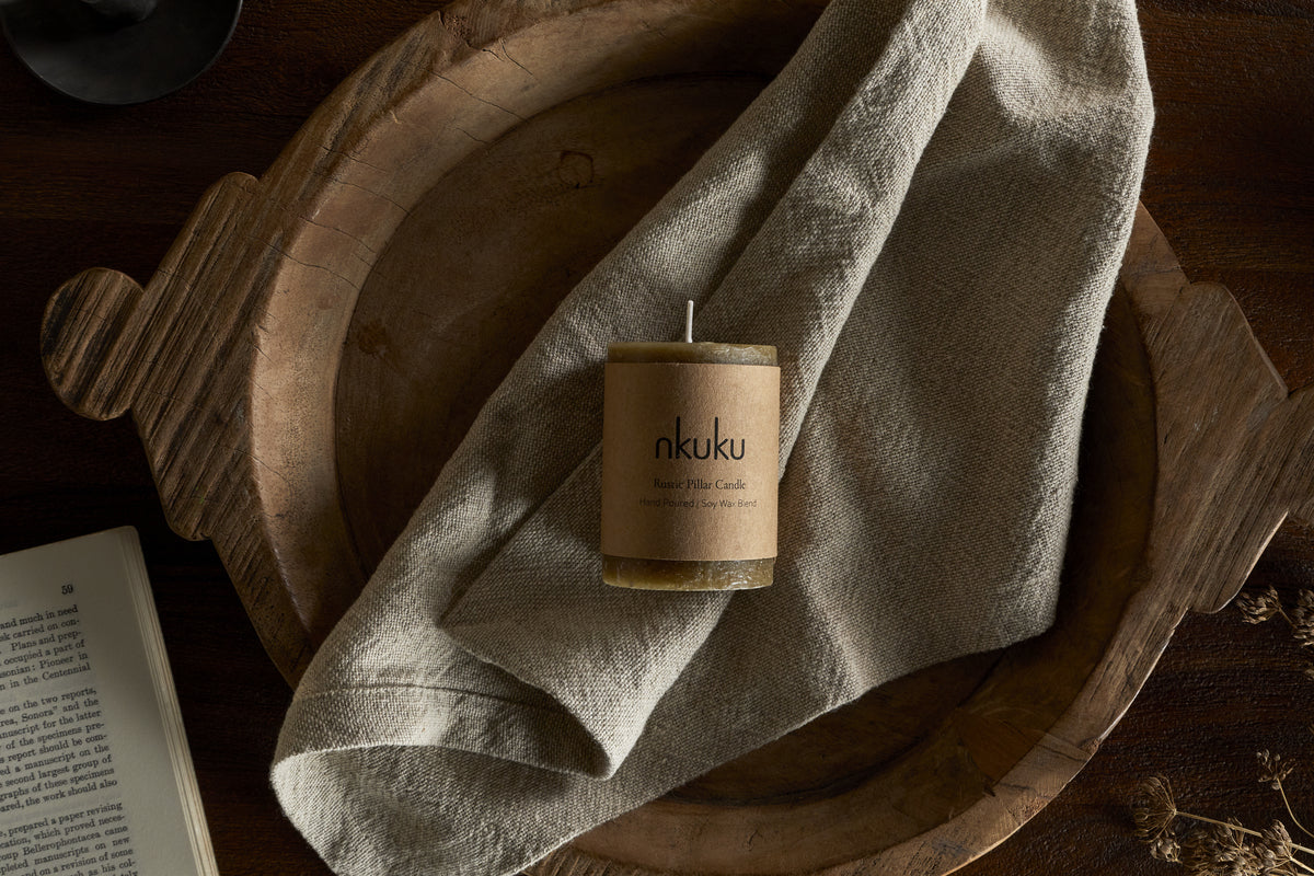 A Rustic Soy Blend Mini Pillar Candle - Olive by nkuku sits on a light beige cloth in a rustic wooden bowl, with part of an open book in the bottom left corner atop a dark wood surface.
