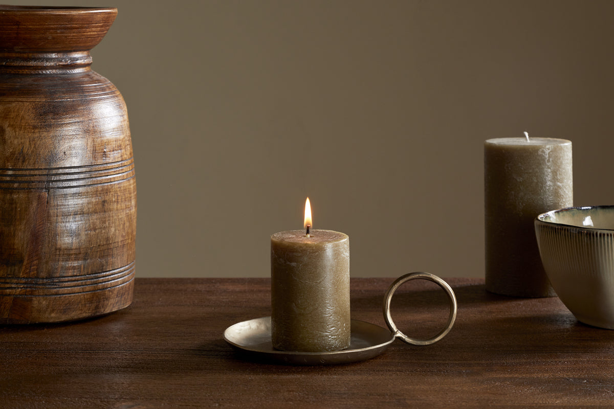 The nkuku Rustic Soy Blend Mini Pillar Candle in Olive glows on a brass handled holder atop a wooden surface, accompanied by a wooden vase, ceramic cup, and an unlit candle, all set against a beige background.