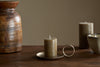 The nkuku Rustic Soy Blend Mini Pillar Candle in Olive sits on a brass holder with handle atop a wooden table, accompanied by a ceramic bowl, another candle, and a textured wooden vase against a plain beige background.