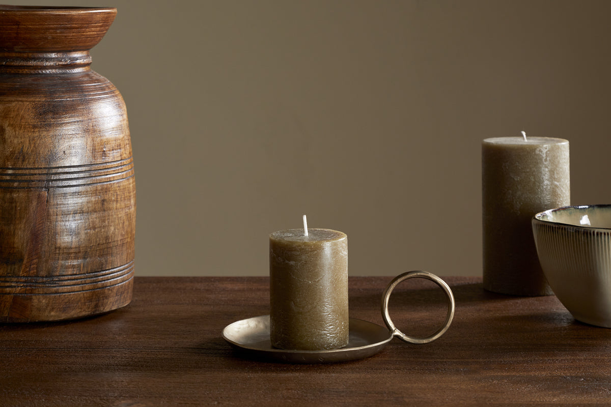 The nkuku Rustic Soy Blend Mini Pillar Candle in Olive sits on a brass holder with handle atop a wooden table, accompanied by a ceramic bowl, another candle, and a textured wooden vase against a plain beige background.