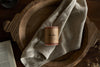 A nkuku Rustic Soy Blend Mini Pillar Candle in a rust finish sits on a folded beige cloth inside a wooden bowl, with an open book nearby on a dark wood surface.