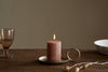 The nkuku Rustic Soy Blend Mini Pillar Candle in rust, with its textured finish, sits on a metal plate to bring charm and warmth to any space.