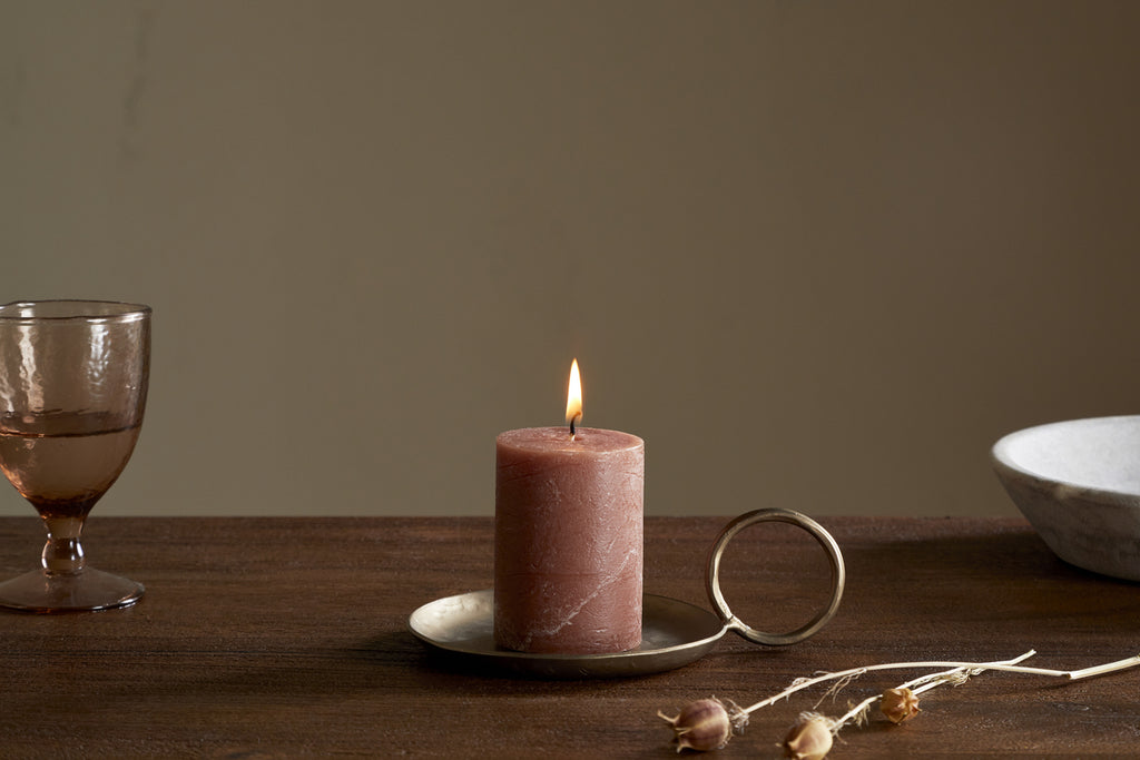 The nkuku Rustic Soy Blend Mini Pillar Candle in rust, with its textured finish, sits on a metal plate to bring charm and warmth to any space.
