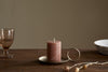 A nkuku Rustic Soy Blend Mini Pillar Candle in Rust with a textured finish sits on a brass handled holder atop a wooden table, beside a pink glass, white bowl, and dried flowers. The softly lit background is plain.