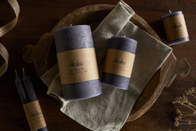 A set of purple textured candles with brown nkuku labels sits on a wooden tray with a folded beige cloth atop a dark wood surface. Nearby are dried flowers, ribbon, and the nkuku Rustic Soy Blend Mini Pillar Candle in Charcoal.