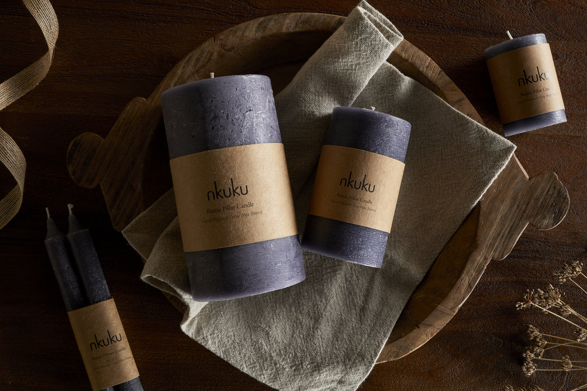 A set of purple textured candles with brown nkuku labels sits on a wooden tray with a folded beige cloth atop a dark wood surface. Nearby are dried flowers, ribbon, and the nkuku Rustic Soy Blend Mini Pillar Candle in Charcoal.