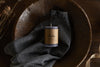 The Rustic Soy Blend Mini Pillar Candle - Charcoal by nkuku, featuring a textured finish, sits on a folded gray cloth in a wooden bowl on a wooden surface, accented by dried flowers.