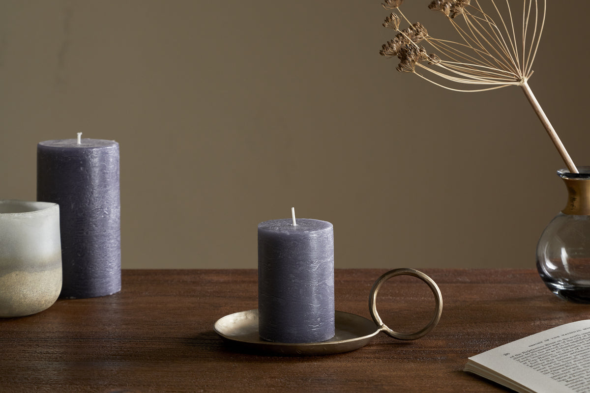 The nkuku Rustic Soy Blend Mini Pillar Candle in Charcoal, featuring a textured finish, sits on the table and adds a charming touch to the setting.