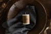 The Rustic Soy Blend Mini Pillar Candle - White by nkuku sits on dark grey cloth in a wooden bowl atop a dark wooden surface, with dried plant stems nearby, showcasing its elegant textured finish.