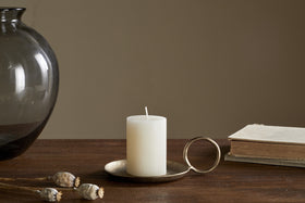 The nkuku Rustic Soy Blend Mini Pillar Candle in white, featuring a textured finish, rests on a brass holder atop a wooden table beside a closed book, dried poppy pods, and a dark glass vase against a beige background.