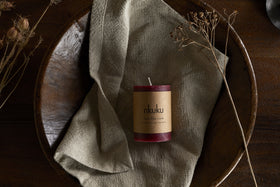 A Rustic Soy Blend Mini Pillar Candle - Dark Cherry by nkuku, with a textured finish, rests on a folded beige cloth in a large wooden bowl, surrounded by dried plants on a dark wooden surface.