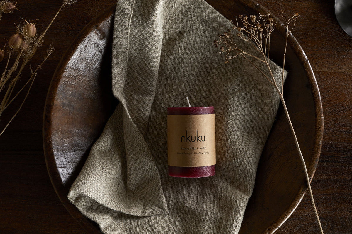 A Rustic Soy Blend Mini Pillar Candle - Dark Cherry by nkuku, with a textured finish, rests on a folded beige cloth in a large wooden bowl, surrounded by dried plants on a dark wooden surface.