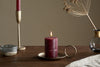 The nkuku Rustic Soy Blend Mini Pillar Candle in Dark Cherry, with a textured finish, sits lit on a brass holder with a ring handle, surrounded by a brass candlestick, small bowl, glass vase of dried flowers, and beige cloth on a wooden table.