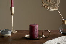 A Rustic Soy Blend Mini Pillar Candle in Dark Cherry by nkuku with a textured finish sits on a brass holder atop a wooden table, beside a gold taper holder with a red candle, a small ceramic bowl, and a glass vase of dried flowers.