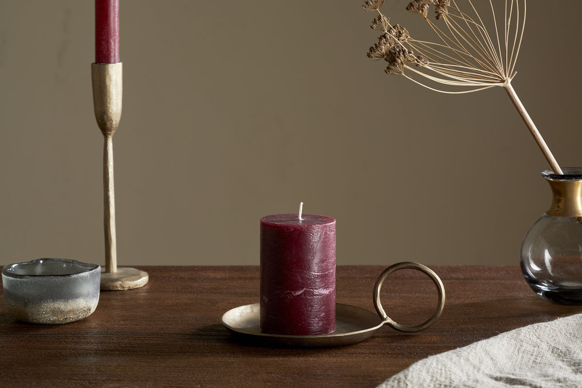 A Rustic Soy Blend Mini Pillar Candle in Dark Cherry by nkuku with a textured finish sits on a brass holder atop a wooden table, beside a gold taper holder with a red candle, a small ceramic bowl, and a glass vase of dried flowers.