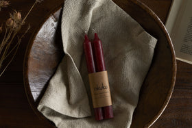 The nkuku Rustic Soy Blend Dinner Candles in Dark Cherry (Set of 2) feature a textured finish and come beautifully wrapped, displayed on a beige cloth in a rustic wooden bowl with dried flowers and an open book on a wooden surface.