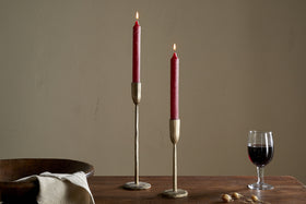 Two Rustic Soy Blend Dinner Candles in Dark Cherry by nkuku sit in brass holders on a wooden table, accompanied by a bowl, folded beige cloth, dried botanicals, and a glass of red wine against a plain beige wall.