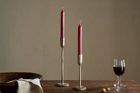 A set of two nkuku Rustic Soy Blend Dinner Candles in Dark Cherry stands on a wooden table beside a glass of red wine, beige napkin, wooden bowl, and dried berries against a plain beige wall.